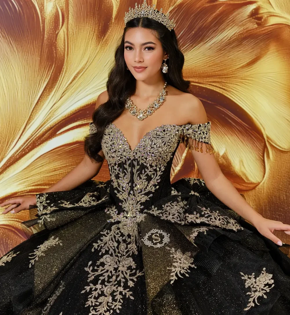 A woman wearing an elaborate black and gold gown with intricate designs, adorned with a tiara and jewelry, posed against a golden floral background.