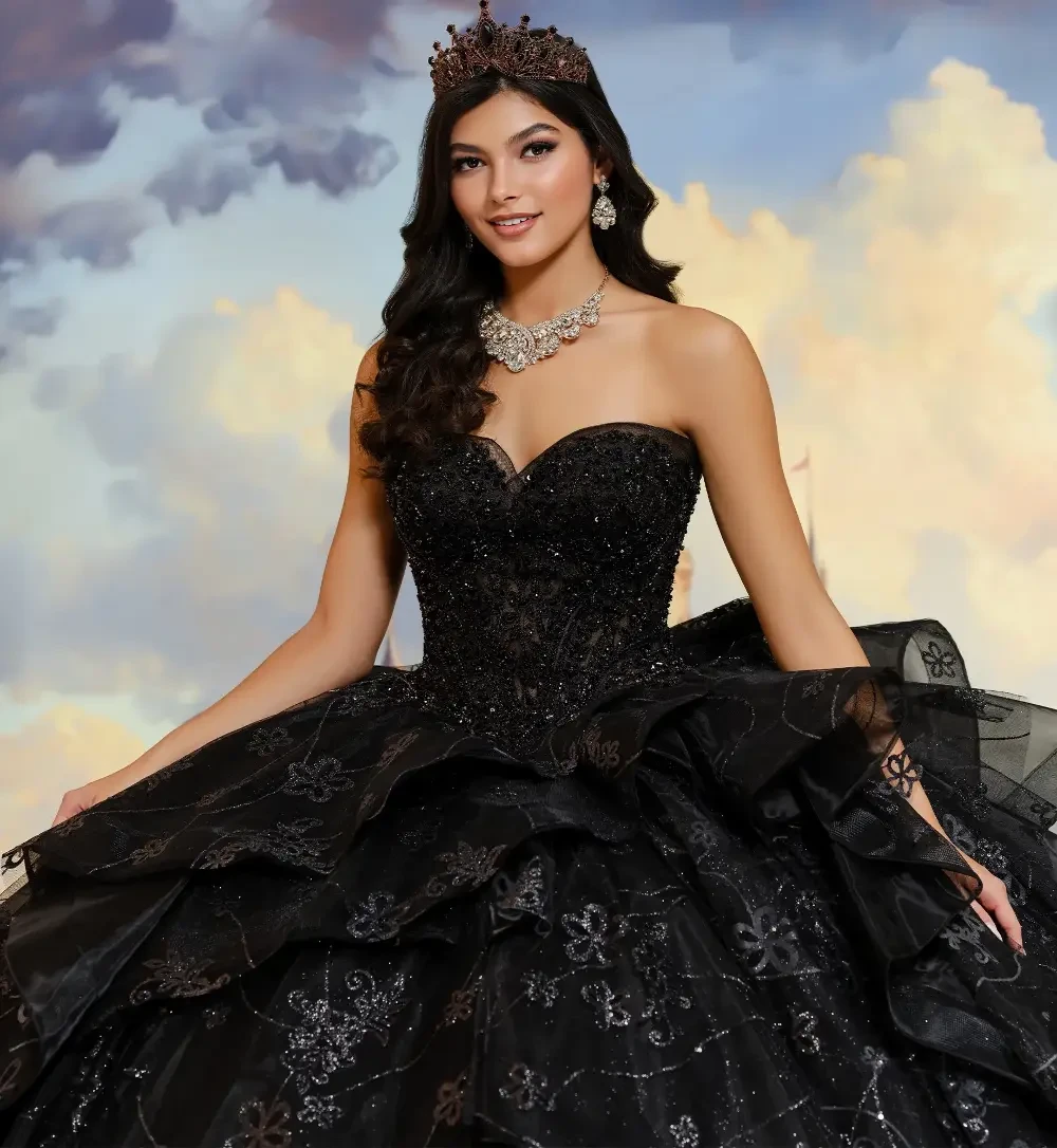 A woman in a black strapless ball gown with ruffles, standing in front of a castle backdrop, wearing a tiara and jewelry.