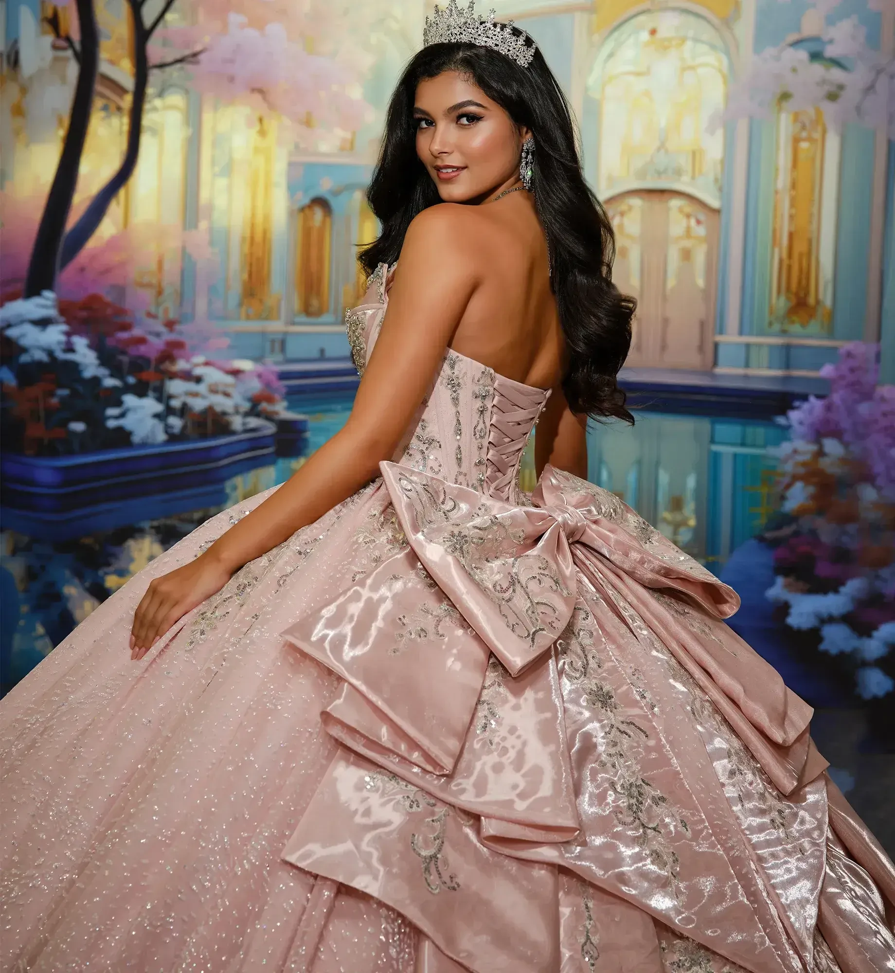 A woman in a sparkling, pastel pink ball gown adorned with a large bow poses gracefully. She wears a tiara and earrings against a dreamy, ornate backdrop.