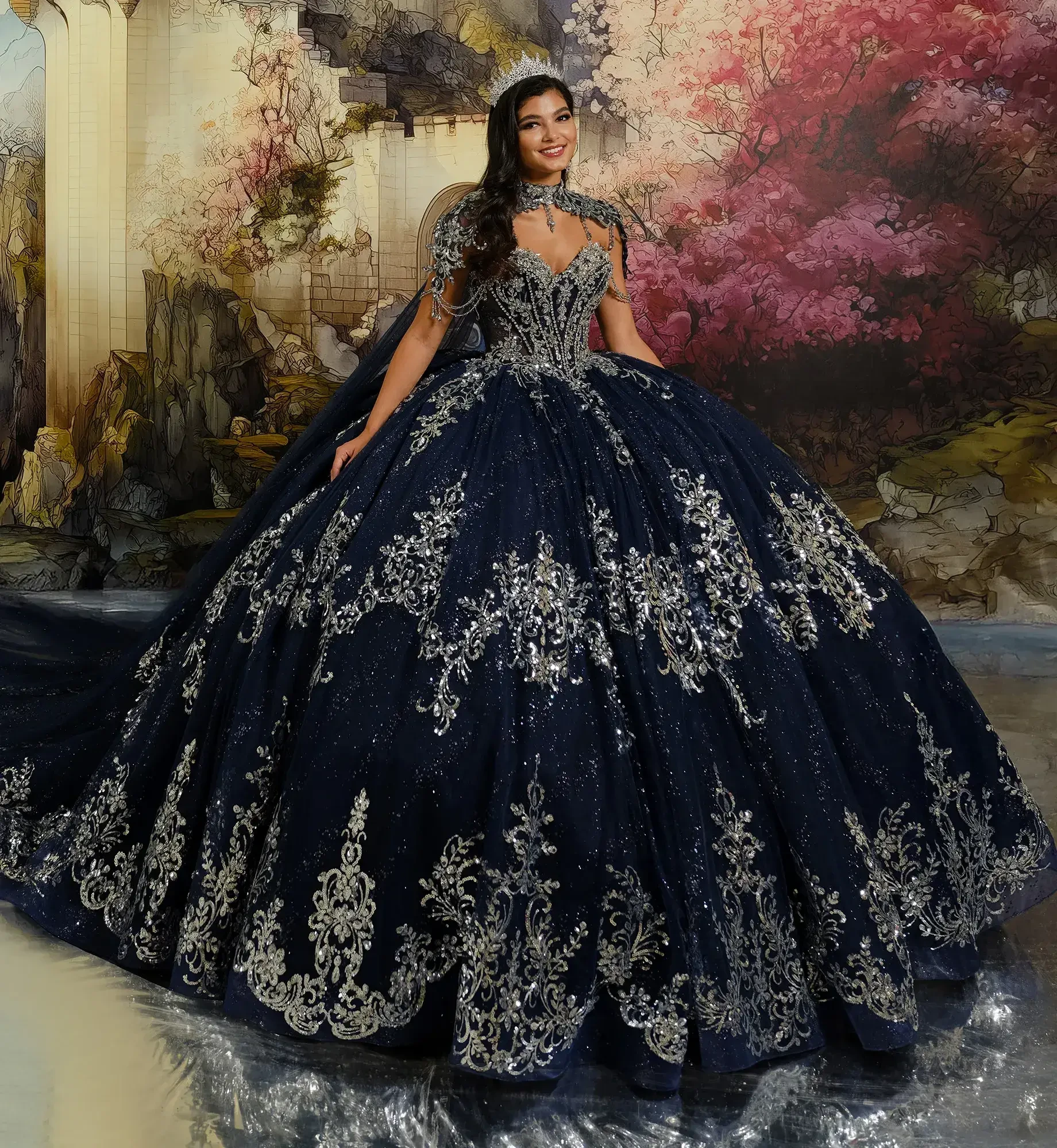 A woman in an ornate, navy ball gown with silver embroidery stands smiling in front of a detailed mural of a castle and blossoms, evoking elegance.
