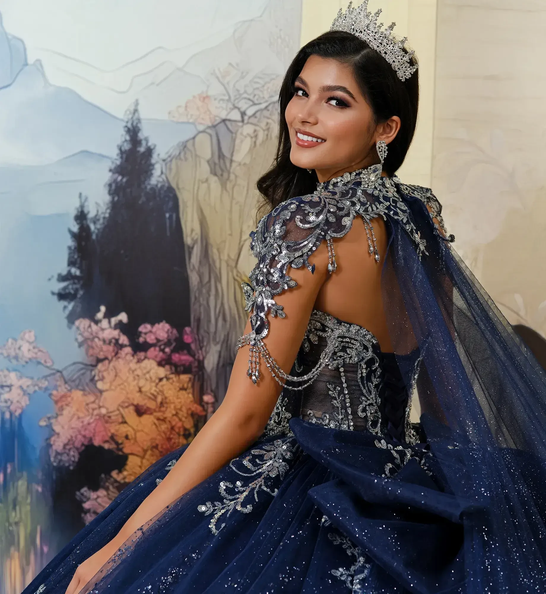 Smiling woman in a sparkling navy blue gown with intricate silver details and a tiara, standing before a colorful nature-themed backdrop.