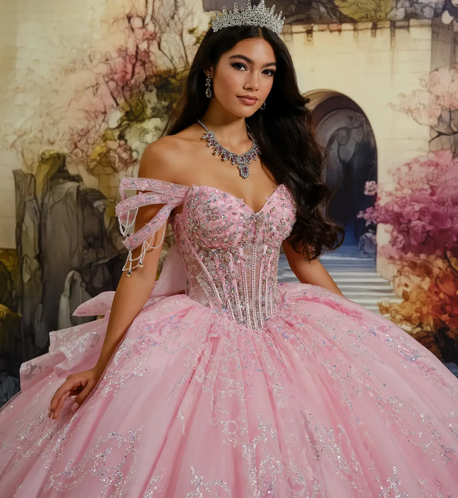 A woman in a sparkling pink ball gown stands smiling in front of a fairytale-like backdrop with arches and vivid autumn trees, exuding elegance.