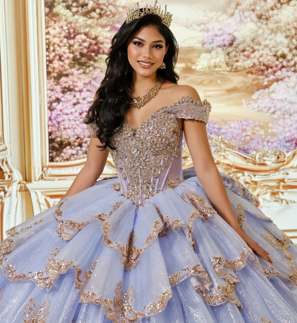 A young woman in a beautiful light purple ball gown with layered ruffles and golden embellishments, standing in an ornate room with floral decorations.